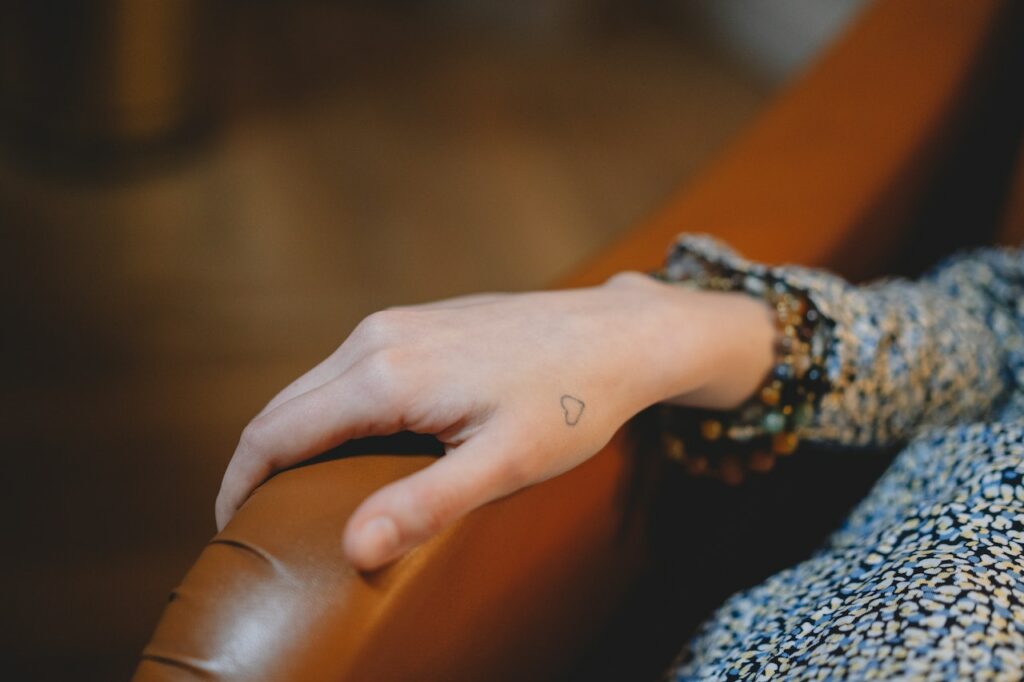A small heart tattoo on the side of a person's hand, sitting on the edge of a couch.