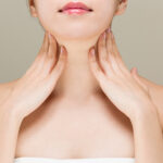 Woman seen from chin down, with hands gracefully placed against her neck.