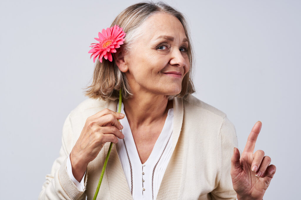 A mature woman smiles as she holds a flower up behind her ear.