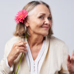 A mature woman smiles as she holds a flower up behind her ear.